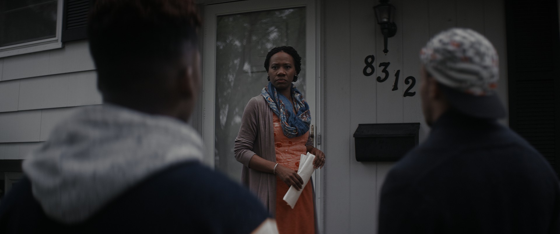 A woman standing on a front porch looks suspiciously at two young men in the foreground.