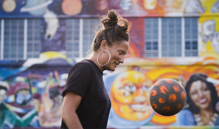 A futbol coach bounces a ball against a backdrop of a colorful mural.